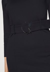 Schwarzer elastischer Gürtel mit einer runden metallischen Schnalle, die eine stilisierte Katzensilhouette zeigt und eine glatte Textur hat. Um einen körpernahen Kleidungsstück befestigt.