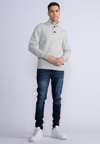Pullover grigio lavorato a maglia con colletto alto, chiusura a due bottoni e orlo a costine. Abbinato a jeans blu scuro e sneaker bianche.