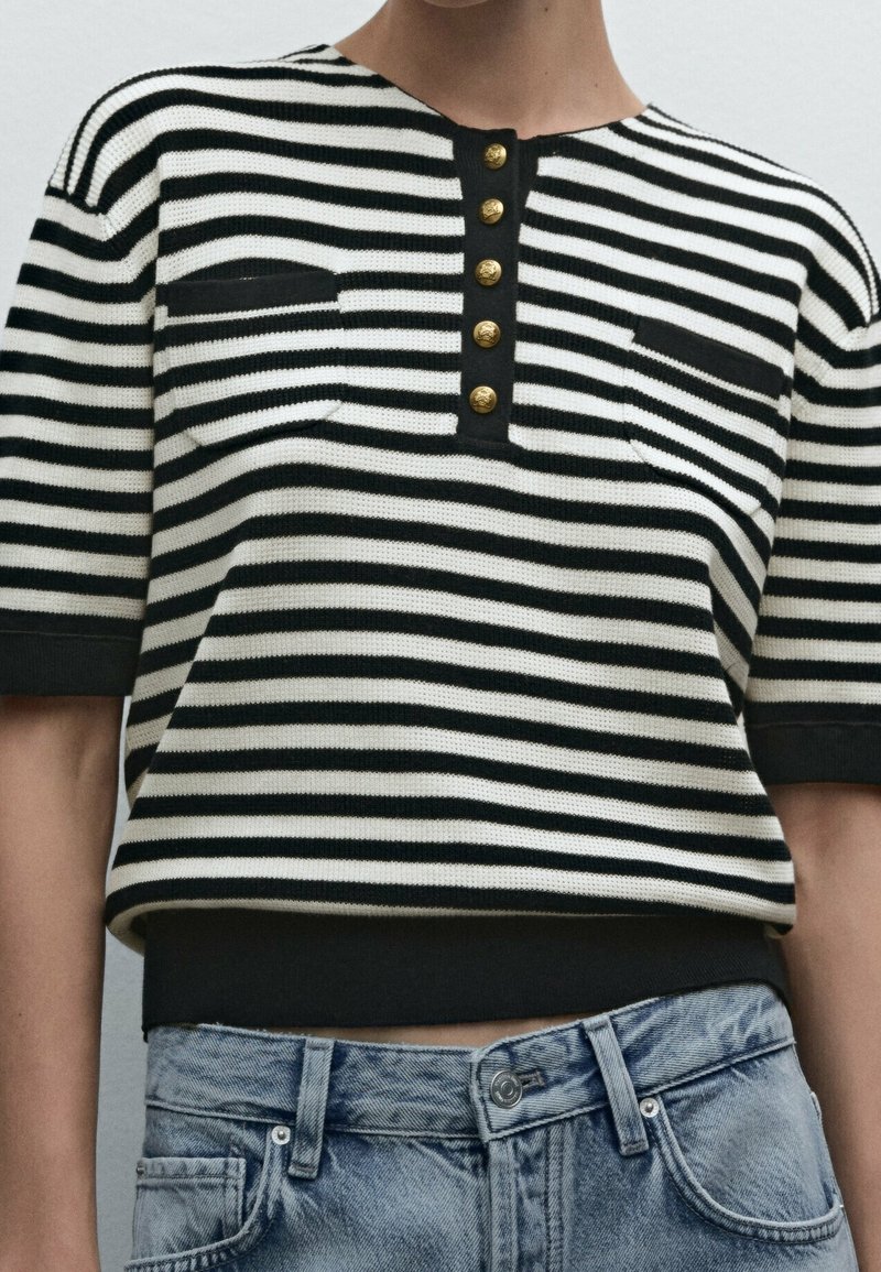 Randig stickad topp i svart och vitt, med rund hals, korta ärmar, fem guldknappar och en framficka.