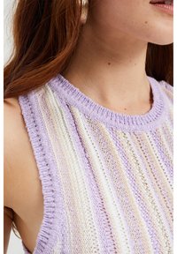 Laventeli ja kerma neule ilman hihoja, jossa on pystysuorat raidat, ribattu makuu ja avoin neuletekstuuri hengittävyyden parantamiseksi.