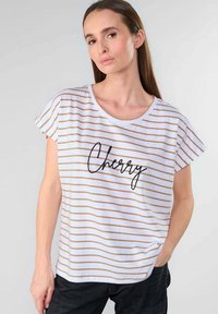 Camiseta blanca con rayas doradas, cuello redondo, mangas cortas, con la palabra "Cherry" en letras cursivas negras en el pecho. Diseño casual.