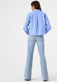 Woman standing with hands in pockets wearing a blue striped shirt and light blue high-waisted flared jeans against a white background.