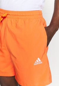 Bañador naranja con cintura elástica y cordón, que presenta un logo blanco de Adidas en la parte inferior izquierda. Tejido suave, de longitud por encima de la rodilla.