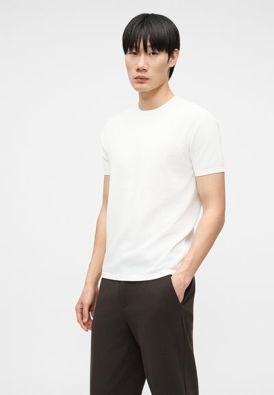 Νεαρός άνδρας με ίσια μαύρα μαλλιά που φοράει ένα απλό λευκό κοντομάνικο μπλουζάκι και σκούρο παντελόνι, στέκεται μπροστά σε ανοιχτό φόντο.