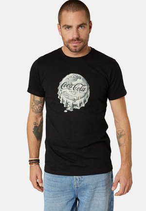 Homme portant un t-shirt noir avec un motif de capsule de bouteille Coca-Cola argentée, un jean bleu clair et des bracelets en perles, debout devant un fond uni.