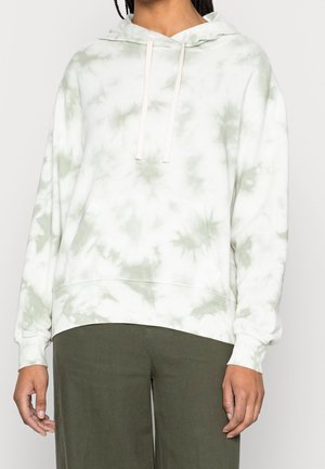 Osoba ubrana w białą i jasnoszarą bluzę z kapturem w stylu tie-dye z troczkami oraz oliwkowozielone spodnie, stojąca z rękami swobodnie opuszczonymi wzdłuż ciała.