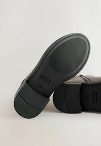 Zwarte rubberen schoenzool met een gestructureerde grip, met een rechthoekig ontwerp, ingedrukte branding en maatmarkeringen, tegen een neutrale achtergrond.