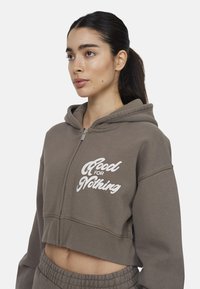Hoodie cropped marrone con zip frontale, caratterizzato da testo bianco "Good for Nothing" sul lato sinistro. Tessuto morbido e vestibilità rilassata.