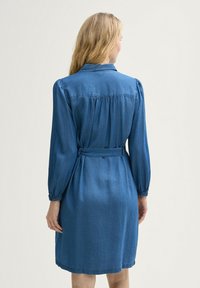 Denim-mekko, vaaleansininen, jossa on kaulus, pitkät puffihuput, kuminauhakääreet ja sidottu vyö vyötäröllä. Selkämyksen laskokset tuovat hienovaraista tekstuuria.
