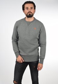 Gray sweatshirt with a crew neck, knit texture, and an orange logo on the chest. Worn with distressed black jeans.