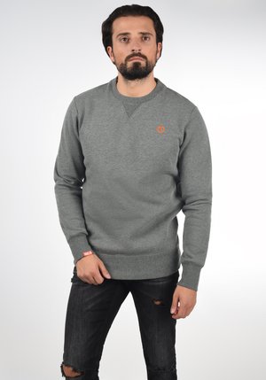 Sudadera gris de cuello redondo, con textura de punto y un logo naranja en el pecho. Llevada con jeans negros desgastados.