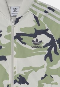 Chaqueta con cierre de Adidas en patrón de camuflaje verde, negro y blanco, con el logotipo de Adidas y tres rayas en el hombro.