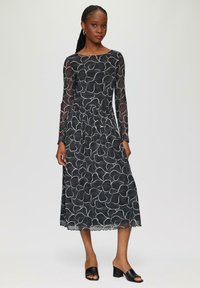 Schwarzes Langarmkleid mit floralem Muster in weißen Linien, tailliert und mit einem gewellten Saum. Getragen mit offenen schwarzen High Heels.