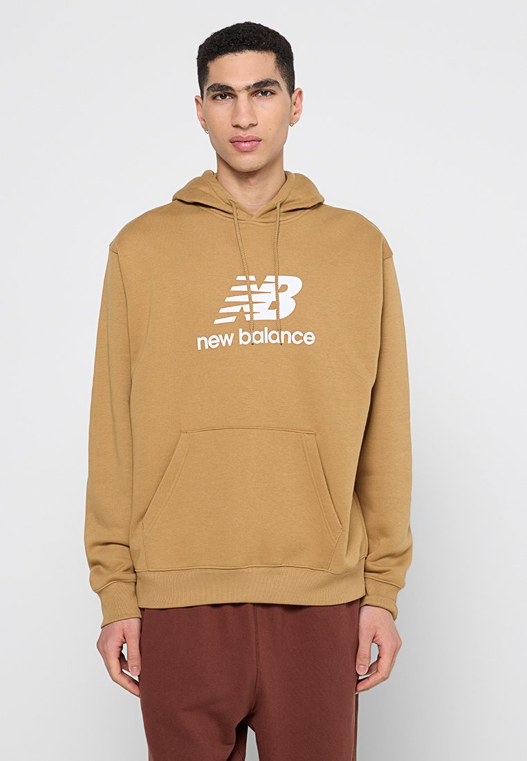 Jeune homme portant un hoodie New Balance couleur camel et un pantalon marron, se tenant contre un fond clair uniforme.