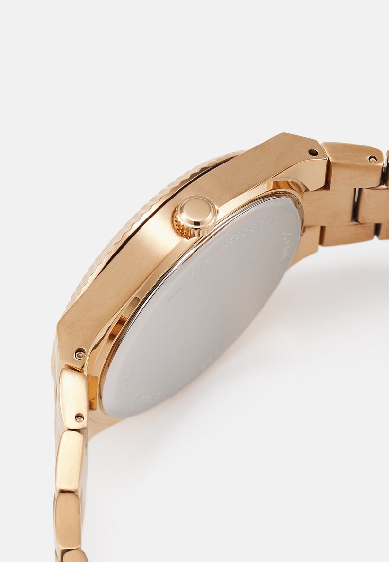 Montre en acier inoxydable plaqué or avec une lunette lisse, une couronne texturée et un bracelet à maillons. Le fond du boîtier est poli avec peu de détails.