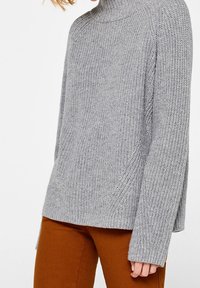 Pull gris tricoté avec un col haut, des manches longues et des fentes latérales, associé à un pantalon marron. La texture est côtelée et douce.