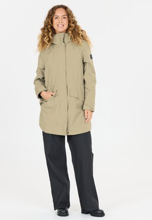Vrouw met krullend haar die lacht, gekleed in een beige parka met hoge kraag, zwarte wijde broek en zwarte schoenen, staand tegen een witte achtergrond.