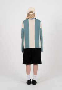 Persoană cu păr lung, purtând un pulover bej cu dungi turcoaz, pantaloni scurți negri, șosete albe, pantofi negri și o șapcă bej, stând cu fața către spate.