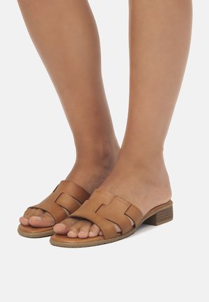 Sandalias deslizantes de cuero marrón claro con un diseño troquelado y tacón bajo. La textura suave y el estilo de punta abierta resaltan el pie.