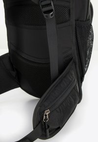 Mochila negra con un panel de malla, correas acolchadas y un pequeño bolsillo con cremallera en el lateral. Tejido texturizado y diseño ergonómico.