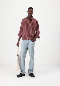 Ein rot-navy kariertes Hemd mit Knöpfen, hellblaue Jeans, schwarze Schuhe und eine cremefarbene Tragetasche, die vor einem weißen Hintergrund steht.