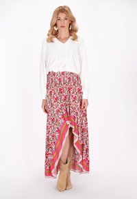 Langer floral Rock mit pinken, grünen und weißen Mustern sowie einem High-Low-Saum. Kombiniert mit einem weißen Langarmoberteil und beigen Stiefeln.