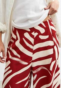 Red and cream patterned trousers featuring abstract brushstroke designs, a lightweight fabric, and a relaxed fit with pockets.
