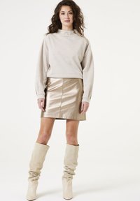 Sudadera gris claro con un cuello alto y un detalle de cremallera dorada, combinada con una falda mini metálica dorada y botas de caña alta en color beige.