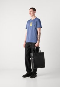 T-shirt de algodão azul com um gráfico amarelo, combinado com calças de ganga escuras. O modelo segura uma bolsa tote de couro preto com alças duplas.