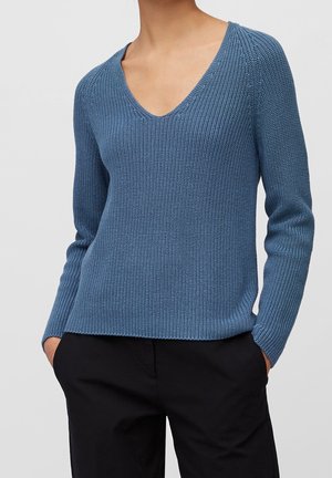 Kvinde iført en blå strikket V-hals sweater med lange ærmer og sorte bukser, hænder i lommerne, foran en ensfarvet grå baggrund.
