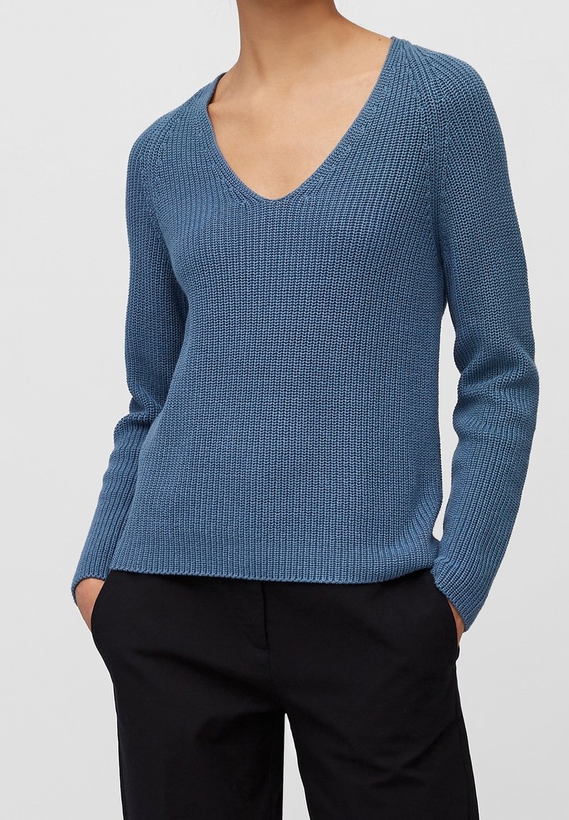 Femme portant un pull en maille bleu à col en V à manches longues et un pantalon noir, les mains dans les poches, sur un fond gris uni.