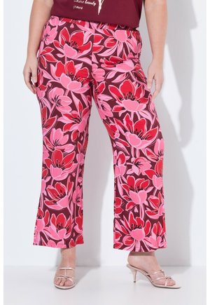 Donna che indossa pantaloni a vita alta e gamba larga con una stampa floreale vivace rossa e rosa, abbinati a sandali beige con tacco e punta aperta, in piedi davanti a uno sfondo bianco.