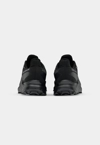 Des chaussures de sport noires présentent une tige en tissu texturé, un bout rond, un col rembourré et une semelle en caoutchouc robuste avec des rainures profondes pour l'adhérence.