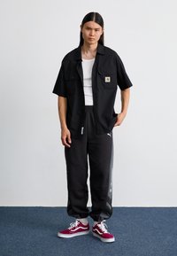 Fekete rövidujjú póló logóval, fehér ujjatlan felső, szürke melegítőnadrág díszítéssel, és piros Vans tornacipő, egyszerű háttér előtt.