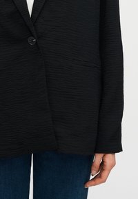 Gros plan d'une personne portant un blazer noir texturé avec un bouton et un jean bleu foncé, main détendue le long du corps.