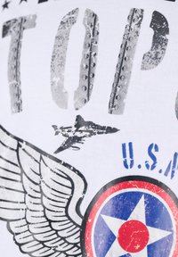 Teksti "TOP" yang tertekan dengan siluet jet militer, grafis sayap yang bergaya, dan lingkaran USA dalam warna merah, putih, dan biru di atas kain putih.