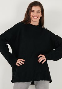 Juodas per didelis megztinis su šoniniu pynimu, aukštu kaklu ir ilgomis rankovėmis. Turi laisvą siluetą ir šoninius plyšius apačioje.