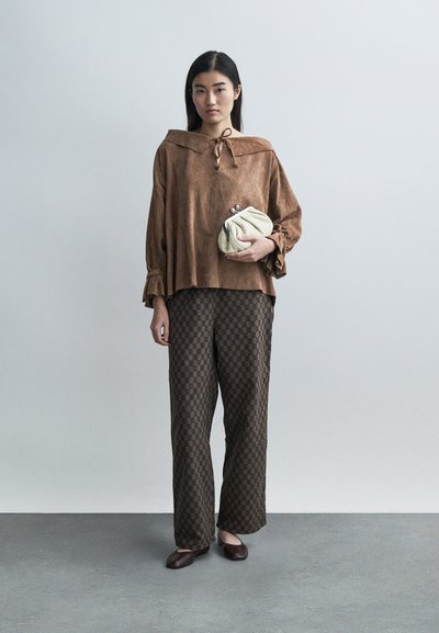 Καφέ φανελένιο τοπ με φαρδιά γιακά και λεπτομέρεια με κορδόνι, συνδυασμένο με καφέ παντελόνι με σχέδιο. Λευκή τσάντα χειρός και σκούρα παπούτσια ολοκληρώνουν το σύνολο.