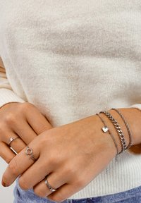 Pulseras de plata en varios diseños: un charm en forma de corazón, una cadena gruesa y una cadena delicada. El anillo presenta una pequeña piedra preciosa. Suéter de crema texturizado.