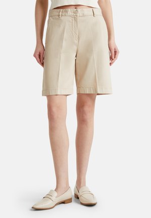 Mujer con pantalones cortos beige a la rodilla y mocasines crema, de pie sobre un fondo blanco liso.
