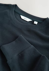 Zwarte crewneck sweatshirt met ribbelstructuur. Voorzien van lange mouwen en een label met de tekst "NEXT" en "REGULAR FIT." Soepele manchet afwerking.