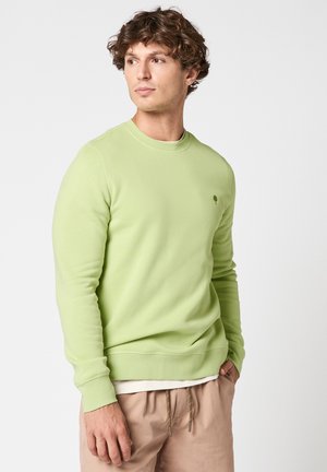 Homme portant un sweat-shirt à manches longues vert clair et un pantalon beige, regardant vers la gauche avec une posture détendue, sur fond clair uni.