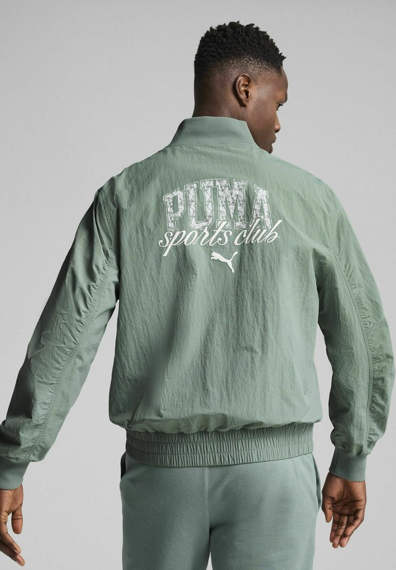 Grøn bomberjakke med elastiske manchetter og talje, der har "PUMA sports club" i hvid tekst på ryggen. Textureret stof, letvægtsdesign.