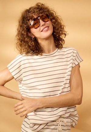 Woman with curly hair wearing large brown sunglasses, striped short-sleeve shirt, and matching drawstring pants, smiling against a beige background.