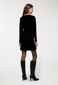 Czarna aksamitna sukienka z długimi rękawami, dopasowaną talią i falbaniastym dołem. Połączona z czarnymi, wysokimi na kolano, szpilkami oraz rajstopami w kropki.