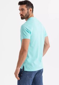 Pikétröja i ljus turkos bomull, kort ärm och slitsar på sidorna. Buren med blå denimjeans, sedd från baksidan.