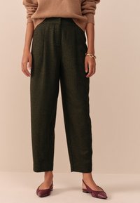 Pantaloni verzi din lână, cu croială lejeră și pliuri frontale, asortați cu pantofi burgundy cu talpă joasă. Modelul poartă un pulover bej.