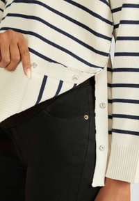Gestreifter Pullover in Creme und Marineblau mit Druckknopfakzenten an der Seite, kombiniert mit schwarzen Jeans, die über ein glänzendes Taschen_detail verfügen.