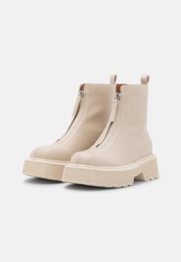 Botas de tobillo de cuero beige con cremallera frontal, suela gruesa estriada y lengüetas en la parte posterior para un fácil calzado. Textura suave en todo el zapato.