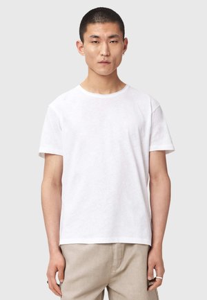 Młody mężczyzna z krótkimi czarnymi włosami, ubrany w białą koszulkę z krótkim rękawem i beżowe spodnie, stojący na tle jasnego, jednolitego tła.